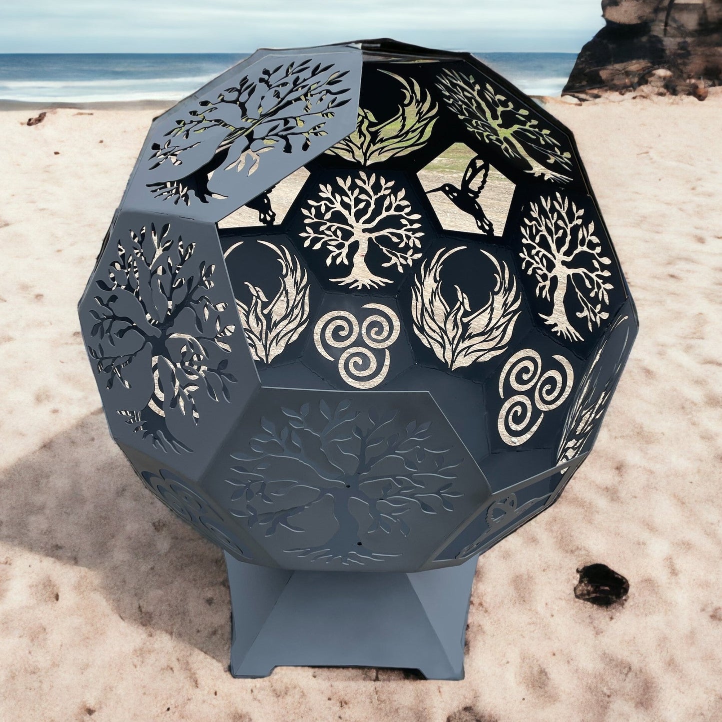 Decorative metal fire pit with earthly designs on a sandy beach.