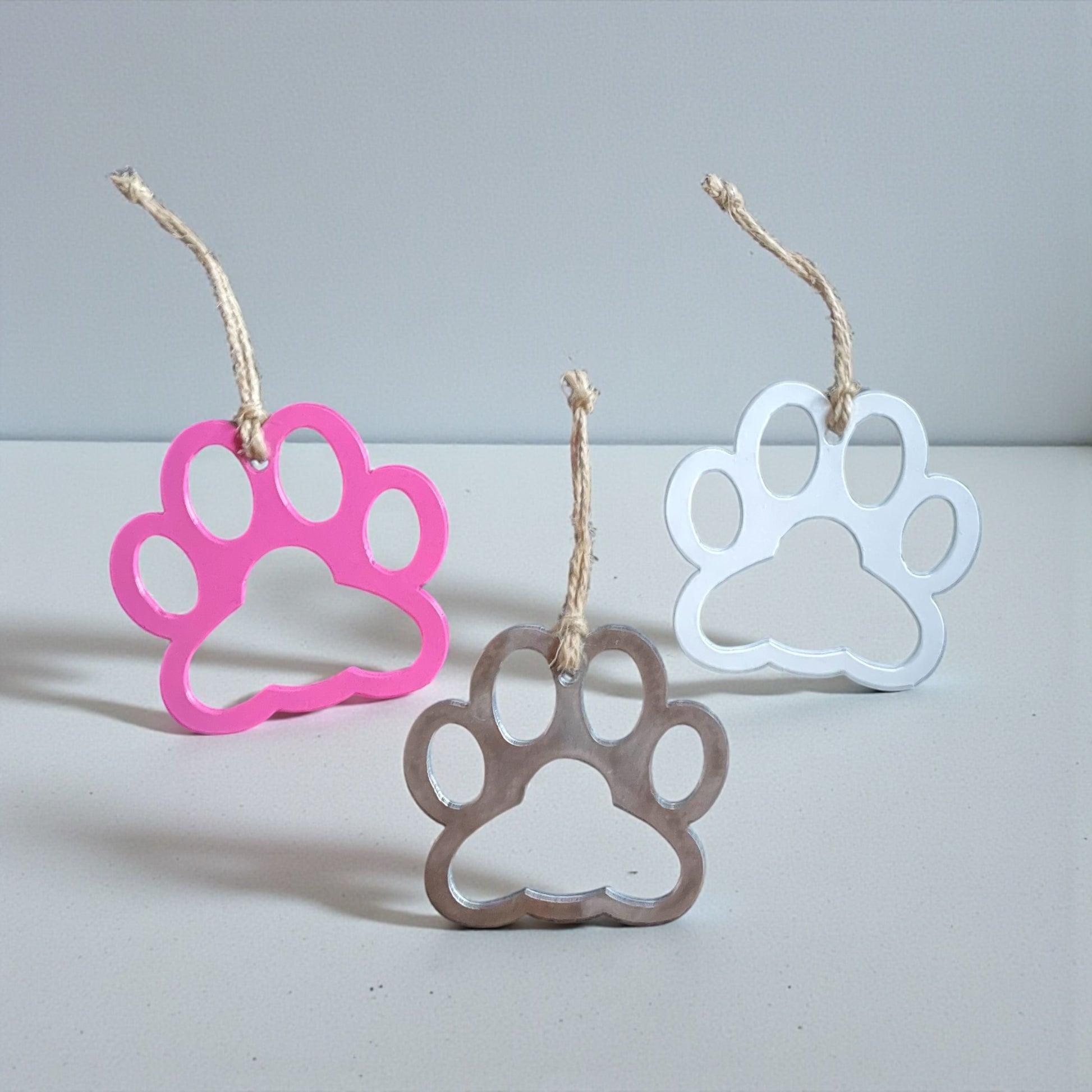 Three paw-shaped Christmas ornaments in pink, brown, and white on a grey white back ground